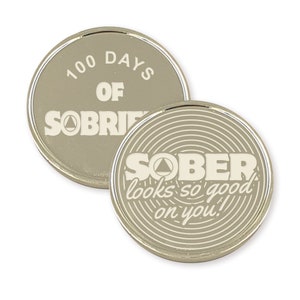 Pu&ograve; includere: Due monete di metallo color argento con un design bianco. La prima moneta recita "100 giorni di sobriet&agrave;" con un simbolo triangolare. La seconda moneta recita "SOBER looks so good on you!" con un simbolo triangolare e un motivo circolare.