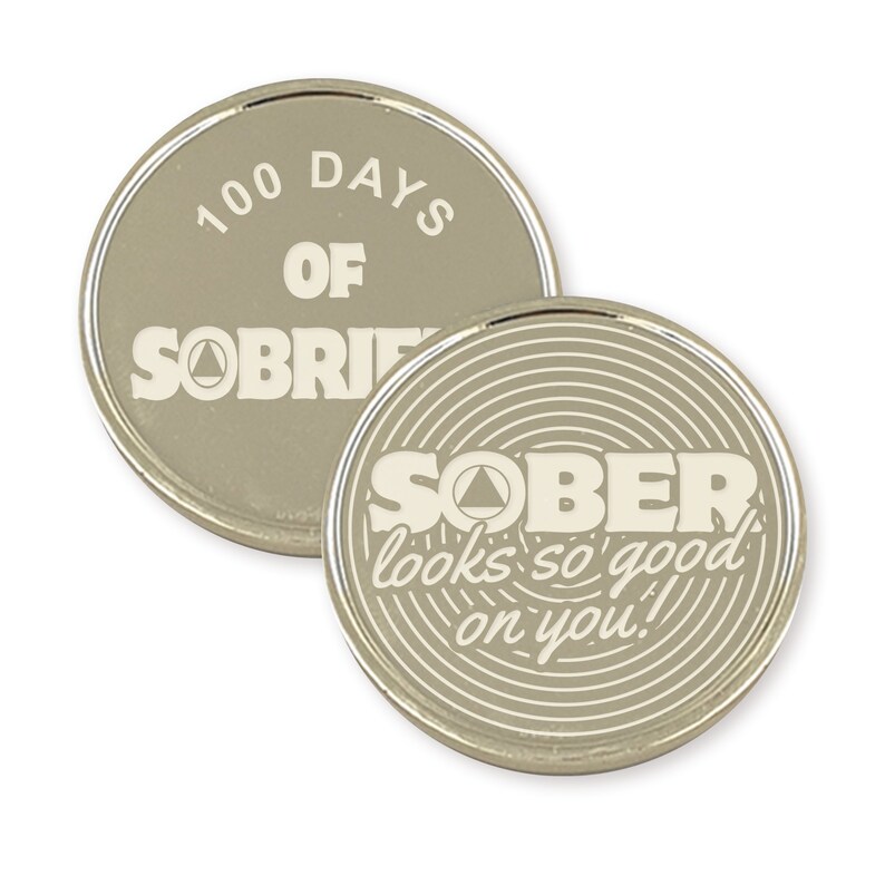 Pu&ograve; includere: Due monete di metallo color argento con un design bianco. La prima moneta recita "100 giorni di sobriet&agrave;" con un simbolo triangolare. La seconda moneta recita "SOBER looks so good on you!" con un simbolo triangolare e un motivo circolare.