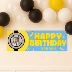 May include: A blue banner with the words "HAPPY BIRTHDAY HANNAH" in yellow. The banner has a photo of a young girl and is decorated with yellow bananas. Black, white, and yellow balloons are in the background.
