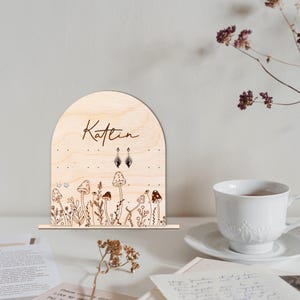 May include: Wooden earring holder with a mushroom and floral design. The holder has a cutout arch at the top and the name "Katlin" is engraved on the front.