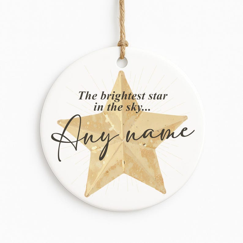 May include: A round, white ceramic ornament with a gold star design. The text "The brightest star in the sky... Any name" is written in black script. A natural twine loop is attached for hanging.