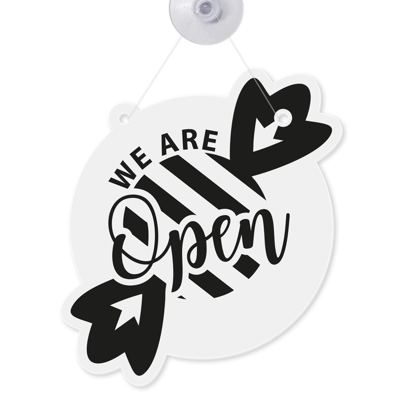 May include: A white and black "We Are Open" sign with a candy-shaped design. The sign is round with the words "We Are" at the top and "Open" in cursive below. The candy design has black stripes and a suction cup for hanging.
