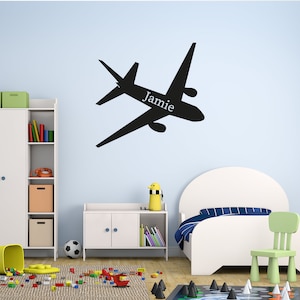 May include: Black airplane silhouette wall decal with the name "Jamie" printed in white. The decal is on a light blue wall above a white bookcase, a white cabinet, and a white bed with a blue and white striped comforter.