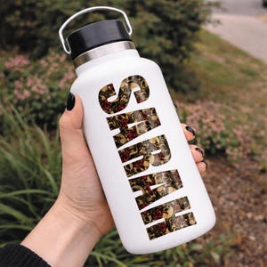May include: White insulated water bottle with a black lid and handle. The word "SARAH" is printed vertically in a pattern of skulls and red roses. The bottle is held against a blurred green background.