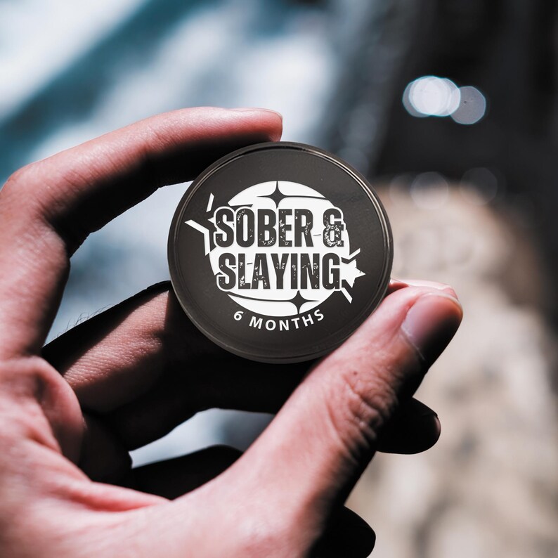 Puede incluir: Moneda redonda en blanco y negro con el texto "SOBER & SLAYING" y "6 MESES" en ella.