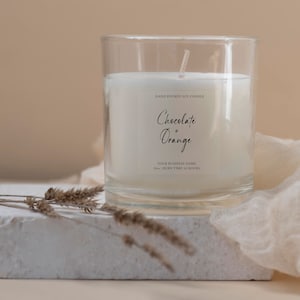 May include: A clear glass candle holder with a white soy candle. The candle has a label that reads "Chocolate + Orange". The candle is on a white surface with dried plants and a beige cloth in the background. The candle is 283g and has a burn time of 10 hours.