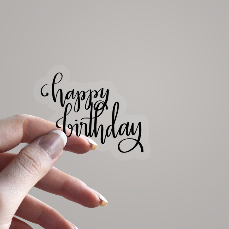 May include: A clear sticker featuring the phrase "happy birthday" in black cursive script. The sticker is held by a hand against a plain background. The lettering is stylish and suitable for birthday celebrations.