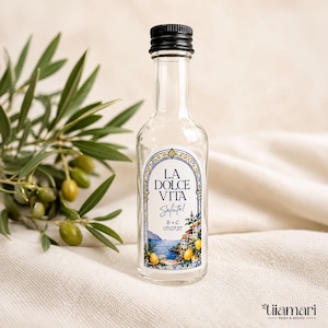 May include: A clear glass miniature bottle with a black screw cap, featuring a decorative label with an arched frame, the text LA DOLCE VITA, Salute!, B + C, 07.07.27, and a coastal scene with lemons, blue water, and white buildings.