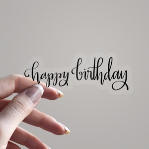May include: A clear sticker featuring the phrase "happy birthday" in black cursive lettering. The sticker is held by a hand with manicured nails against a neutral backdrop. The script has a graceful, flowing design.
