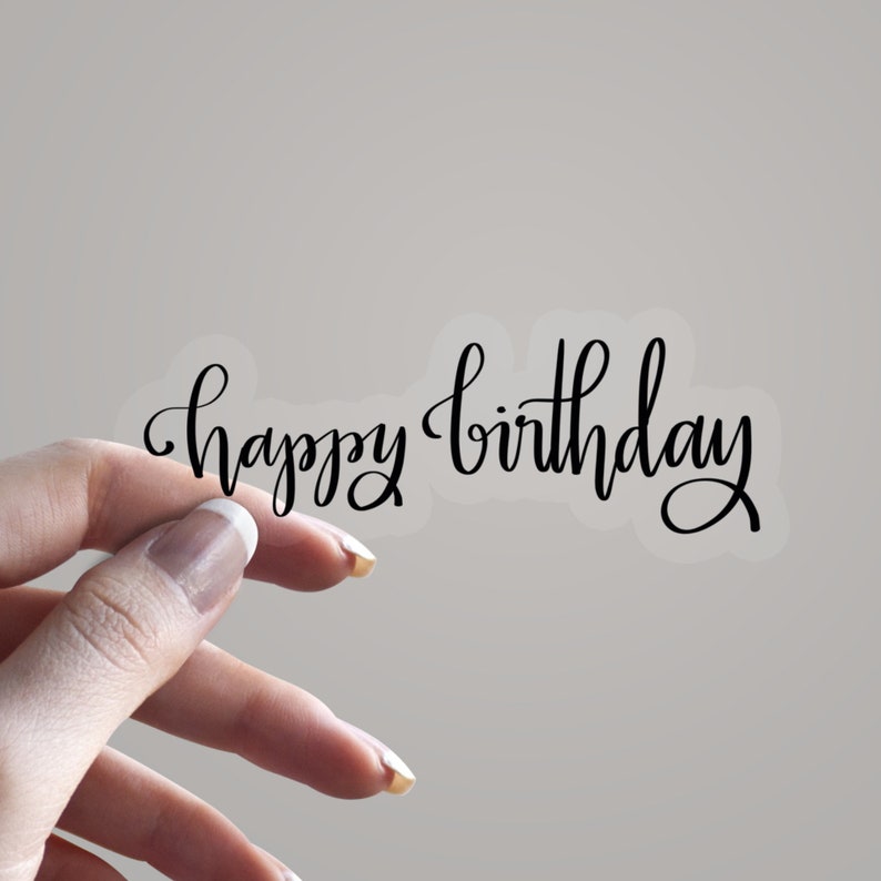 May include: A clear sticker featuring the phrase "happy birthday" in black cursive lettering. The sticker is held by a hand with manicured nails against a neutral backdrop. The script has a graceful, flowing design.