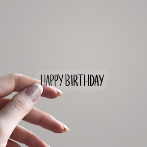 May include: A transparent sticker with the text "HAPPY BIRTHDAY" in a black, handwritten font. The sticker is held by a hand, set against a plain background.