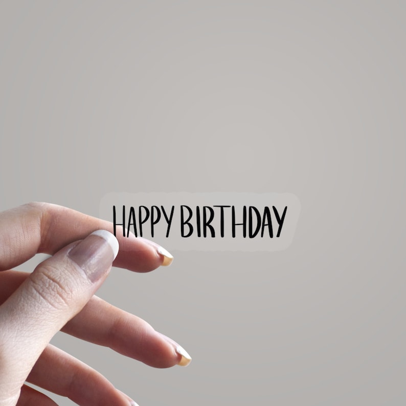 May include: A transparent sticker with the text "HAPPY BIRTHDAY" in a black, handwritten font. The sticker is held by a hand, set against a plain background.