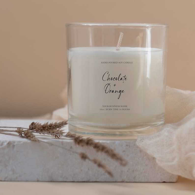 May include: A clear glass candle holder with a white soy candle. The candle has a label that reads "Chocolate + Orange". The candle is on a white surface with dried plants and a beige cloth in the background. The candle is 283g and has a burn time of 10 hours.