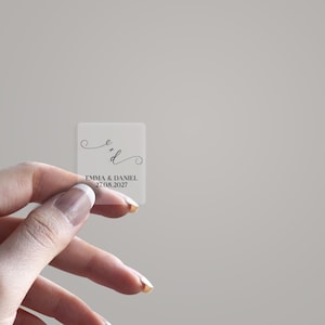 May include: A small, clear, rectangular sticker held by a hand. The sticker features the initials "e" and "d" in a delicate, cursive font, along with the names "EMMA & DANIEL" and the date "27.08.2027" in a simple, sans-serif font.