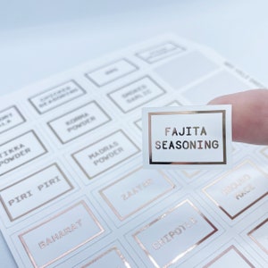 May include: A sheet of white spice labels with rose gold trim. One label is held up, displaying "FAJITA SEASONING" in rose gold lettering. Other labels include "MADRAS POWDER", "CHIPOTLE", and "ZAATAR".