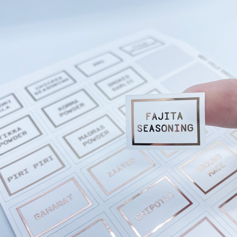 May include: A sheet of white spice labels with rose gold trim. One label is held up, displaying "FAJITA SEASONING" in rose gold lettering. Other labels include "MADRAS POWDER", "CHIPOTLE", and "ZAATAR".