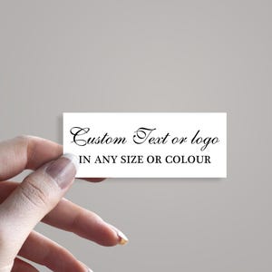 Puede incluir: Una tarjeta rectangular blanca con el texto "Custom Text or logo IN ANY SIZE OR COLOUR" en escritura negra. La tarjeta está sostenida por una mano sobre un fondo gris claro. El texto sugiere opciones de personalización.