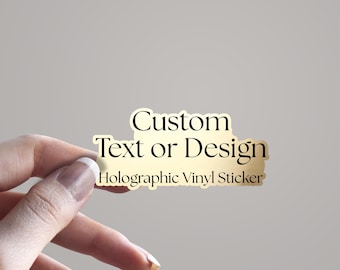 May include: A gold holographic vinyl sticker with the text "Custom Text or Design" and "Holographic Vinyl Sticker" in black. The sticker is held by a hand against a neutral background.