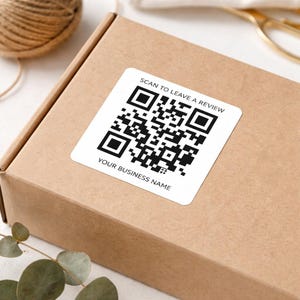 Könnte beinhalten: Ein quadratischer weißer Aufkleber mit einem schwarzen QR-Code und dem Text "SCAN TO LEAVE A REVIEW" und "YOUR BUSINESS NAME" ist auf einer braunen Pappschachtel angebracht. Garn, Grünzeug und eine goldfarbene Schere sind ebenfalls zu sehen.