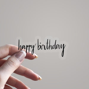 May include: A transparent sticker featuring the phrase "happy birthday" in black, handwritten-style lettering. The sticker is held by a hand against a plain backdrop.