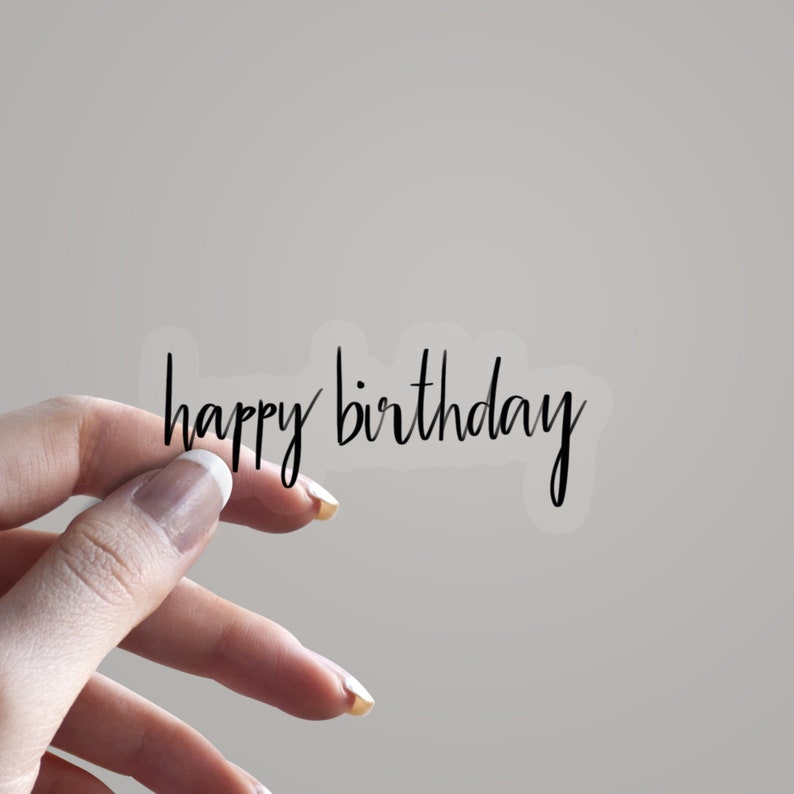 May include: A transparent sticker featuring the phrase "happy birthday" in black, handwritten-style lettering. The sticker is held by a hand against a plain backdrop.