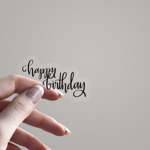 May include: A transparent sticker featuring the phrase "happy birthday" in black cursive lettering. The sticker is held up by a hand. Ideal for adding a personal touch to gifts or decorations.