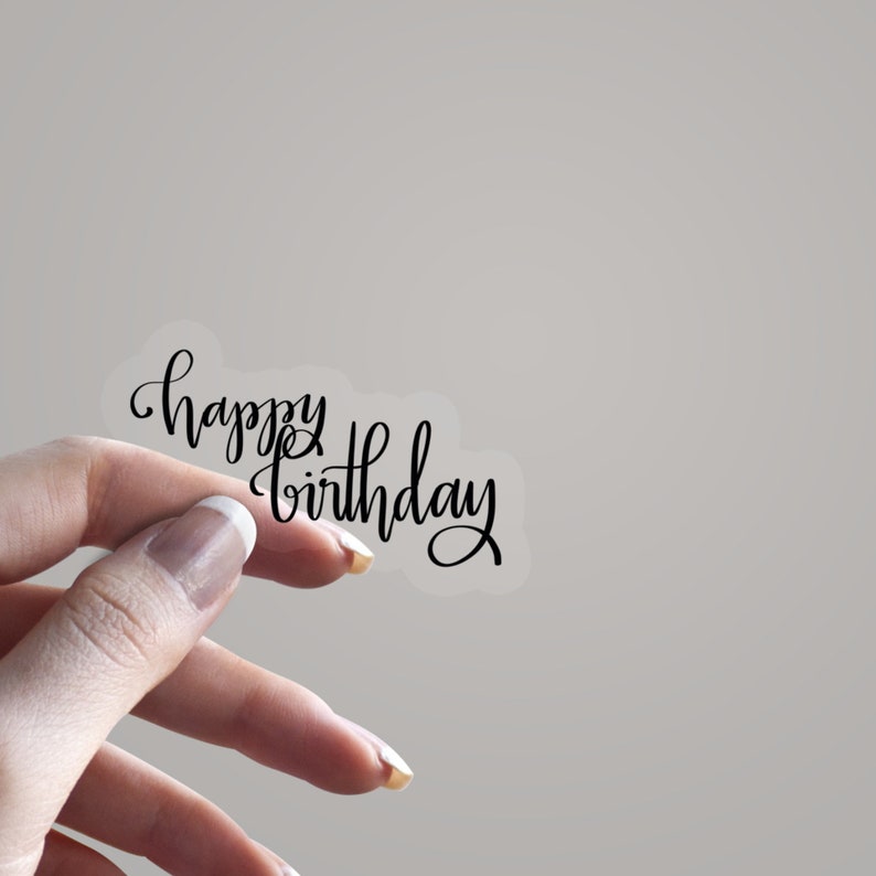 May include: A transparent sticker featuring the phrase "happy birthday" in black cursive lettering. The sticker is held up by a hand. Ideal for adding a personal touch to gifts or decorations.