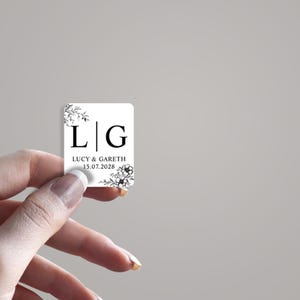 May include: A white square wedding favour tag with black floral designs and the initials "L" and "G" separated by a vertical line. The text "Lucy & Gareth 15.07.2028" is printed below the initials.