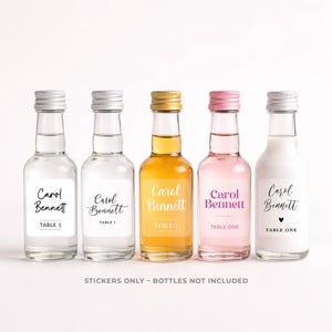 May include: Five miniature glass bottles with silver, gold, and pink screw caps, each featuring a custom rectangular label with the name Carol Bennett and table number text. The bottles contain clear, amber, pink, and opaque white liquids.