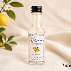 May include: A clear glass miniature bottle with a black screw cap, featuring a rectangular label with a blue and yellow decorative border. The label reads Cheers TO FOREVER, LIMONCELLO, BEA & TIMOTHY, and 27.06.2028, with a small lemon illustration.