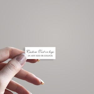 Puede incluir: Una pequeña etiqueta rectangular blanca con el texto "Custom Text or logo IN ANY SIZE OR COLOUR" en escritura negra. La etiqueta se sostiene entre los dedos sobre un fondo gris neutro.