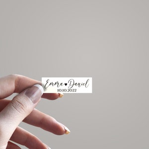 May include: White rectangular wedding favor tag with black calligraphy text that reads "Emma &hearts; Daniel 10.10.2022".