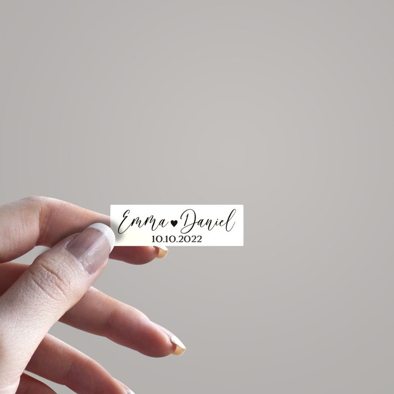 May include: White rectangular wedding favor tag with black calligraphy text that reads "Emma &hearts; Daniel 10.10.2022".
