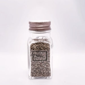 May include: A clear glass spice jar with a metal lid, filled with dried basil. The label on the front reads "BASIL" in a black font. The jar is rectangular with rounded corners, and the contents are a brownish-green color.