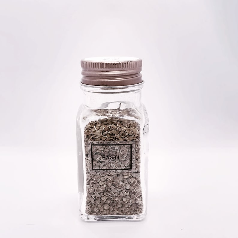 May include: A clear glass spice jar with a metal lid, filled with dried basil. The label on the front reads "BASIL" in a black font. The jar is rectangular with rounded corners, and the contents are a brownish-green color.