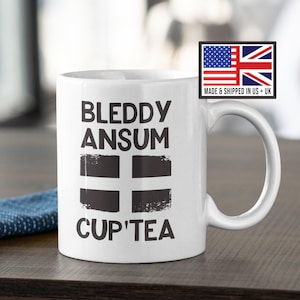 Puede incluir: Taza de cerámica blanca con las palabras "BLEDDY ANSUM CUP'TEA" en negro y un diseño de cruz en blanco y negro. La taza tiene un asa curva y una pegatina con las banderas de EE. UU. y Reino Unido.