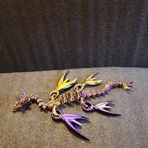 Puede incluir: Una figurita de dragón de color púrpura y dorado con un cuerpo segmentado y alas. El dragón está acostado de lado con la cabeza girada hacia la izquierda.