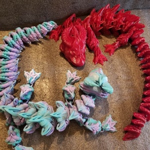 May include: Two 3D printed dragon figures, one red and one blue and purple. The red dragon is lying down with its head facing the viewer. The blue and purple dragon is curled up in a circle.