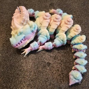 May include: A colorful, articulated dragon figurine in pastel shades of pink, blue, and yellow. The dragon has a detailed head with sharp teeth and a segmented body. The toy is made of plastic and appears to be 3D printed.