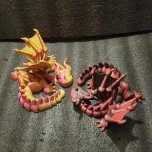 May include: Two small, 3D printed dragon figurines. One is a yellow and pink dragon with its wings spread out. The other is a red dragon with its wings folded.