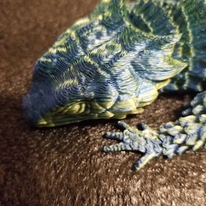 May include: Close-up of a blue and green 3D printed lizard figurine. The lizard has a textured, scaly surface and is posed with its head down and its front leg extended.