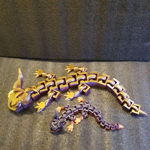 May include: Two articulated lizard sculptures. The larger lizard is gold and purple, with articulated segments and small feet. The smaller lizard is dark purple and orange. Both are on a dark gray surface.