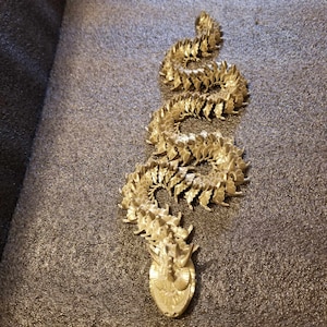 May include: A gold-colored metal sculpture of a snake with a detailed, textured surface. The snake is curled in a spiral shape, with its head at the bottom of the sculpture.