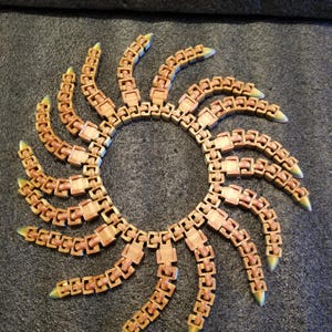 May include: A circular, segmented sculpture with a sunburst design. The sculpture is made of light brown, interlocking pieces with a slight wood grain appearance. The tips of each segment are tipped with a pale green color.