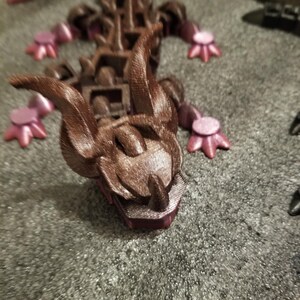 May include: A brown and purple 3D-printed dragon figurine. The dragon has a detailed head with horns and a textured body. The feet are a lighter purple color. The figurine is on a gray surface.