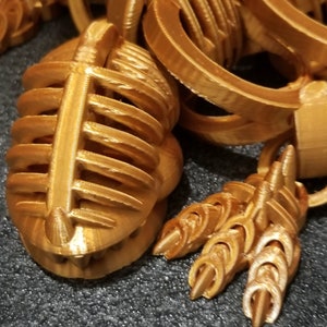 以下が含まれることがあります： 金色の3Dプリントされたオブジェクトで、ケージのようなデザインと3つの爪のような付属物が付いています