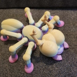 Peut inclure: Un jouet araignée imprimé en 3D aux couleurs pastel. L'araignée a un corps arrondi et des pattes segmentées, avec une couleur beige clair sur le corps et les pattes, et des accents roses et bleus. Le jouet est conçu avec une surface texturée détaillée.
