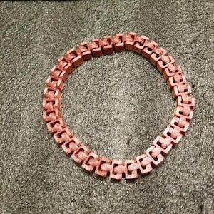 May include: A circular, reddish-brown bracelet composed of interlocking rectangular links. The bracelet is made of a textured material and is displayed against a dark, speckled background. The design is geometric and modern.