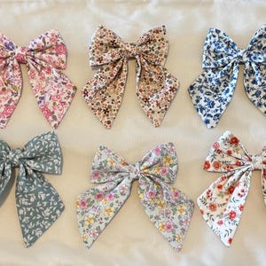 May include: Six fabric hair bows in various floral patterns. The bows feature a variety of colors, including pink, blue, and white, with detailed floral designs. Each bow has long, flowing tails.