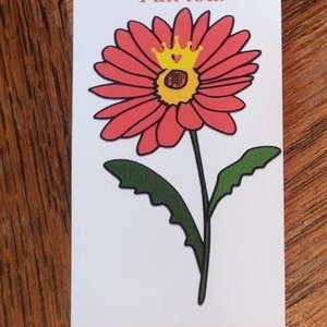 May include: A pink flower with a yellow center and a gold crown on top. The flower is on a white background with the text "Queen of the Fall tour".